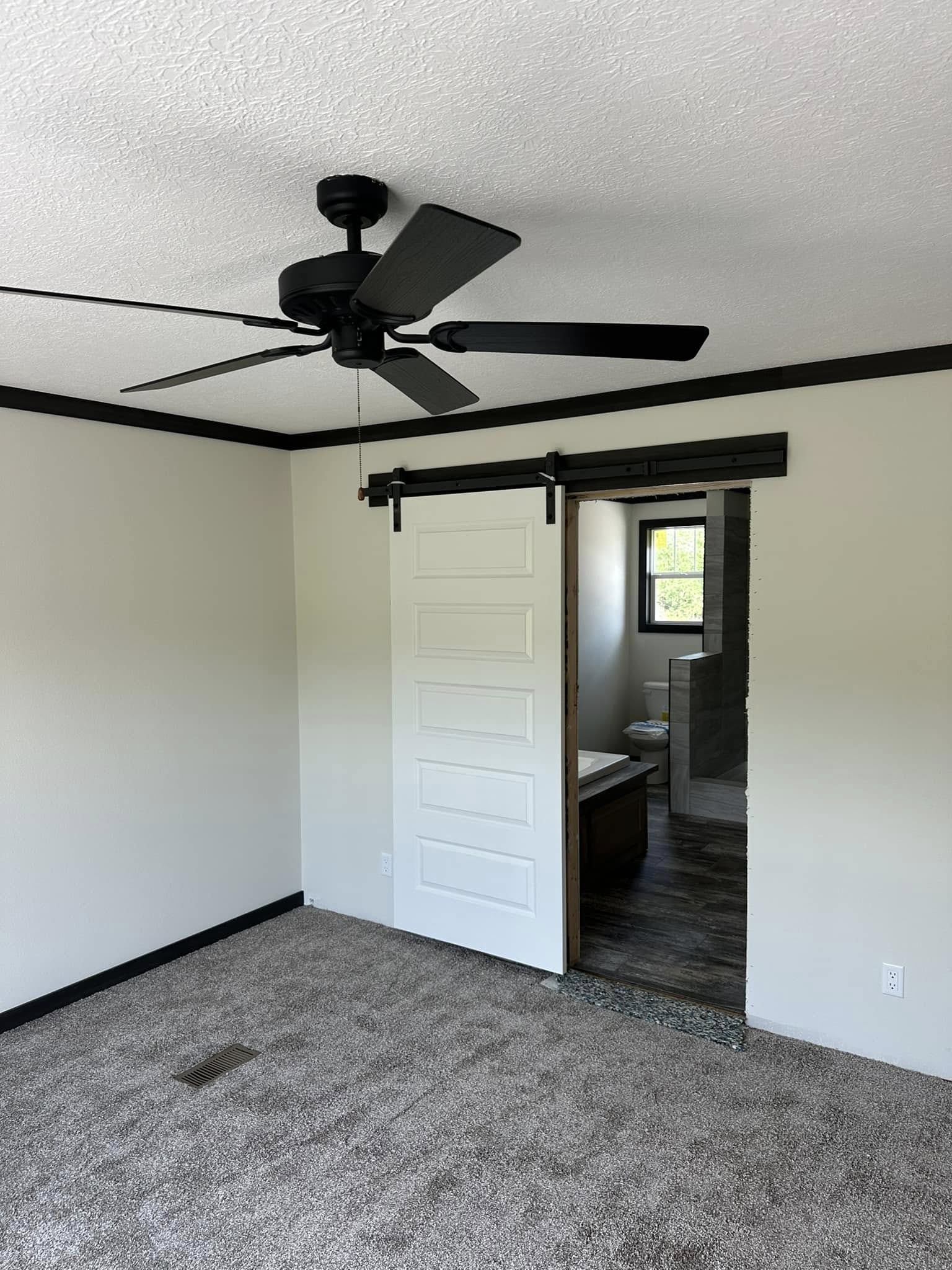 Spacious room with beige walls, brown carpet, a black ceiling fan, and a sliding white barn door leading to a bathroom, creating a modern, minimalist feel.