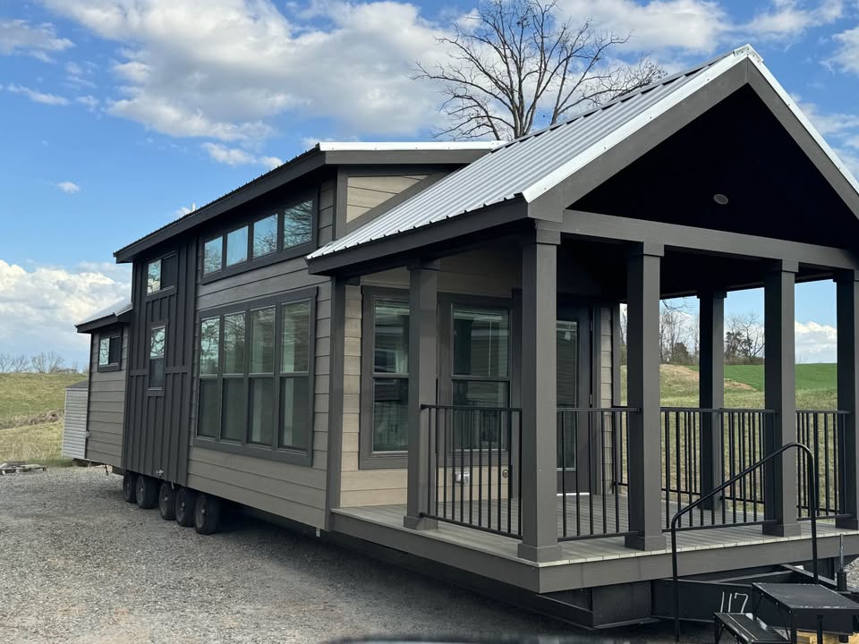 Modern tiny house on wheels with a dark exterior and large windows. It has a metal roof and a covered porch, set against a blue sky and grassy landscape.
