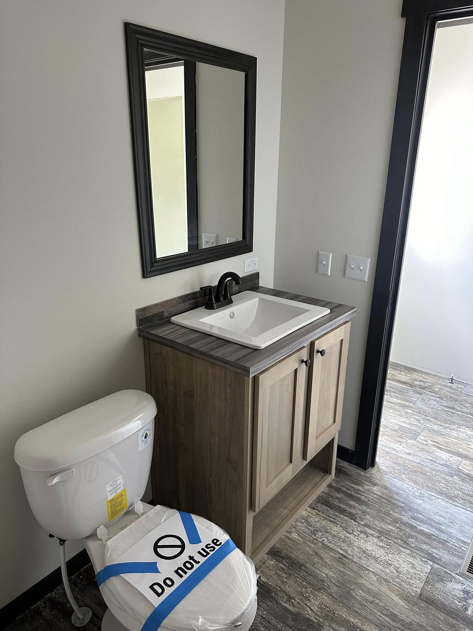 Bathroom with a wooden vanity and sink under a mirror, a toilet covered with a "Do not use" sign, set against light-colored walls and tile floor.