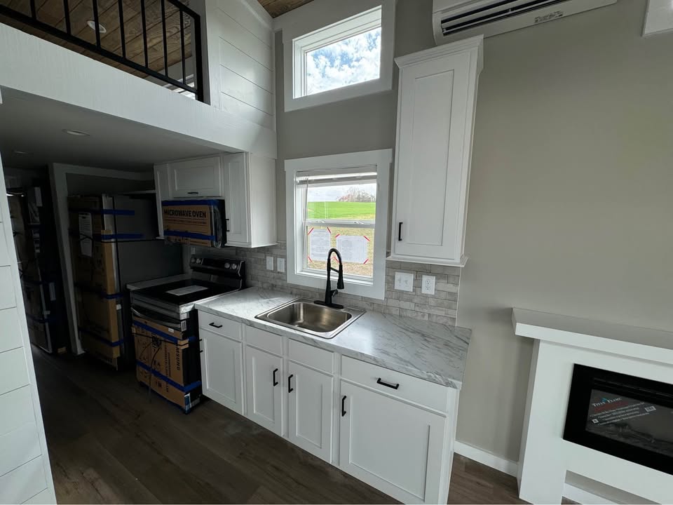 Compact kitchen with white cabinets, a marble countertop, and a sink beneath a window. New appliances are boxed. Calm, modern atmosphere.