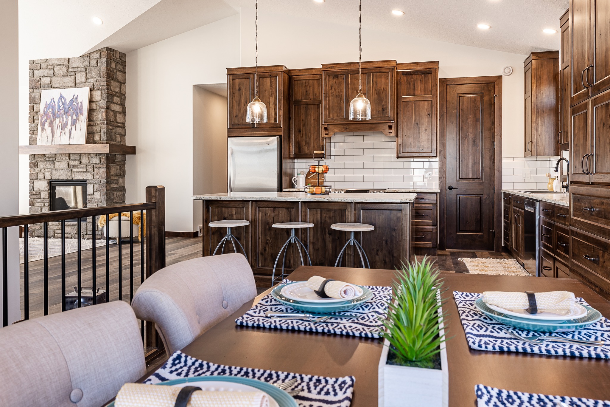 Spacious kitchen with wooden cabinets and island, pendant lights, and a stone fireplace. Dining table set with patterned placemats and rolled napkins.