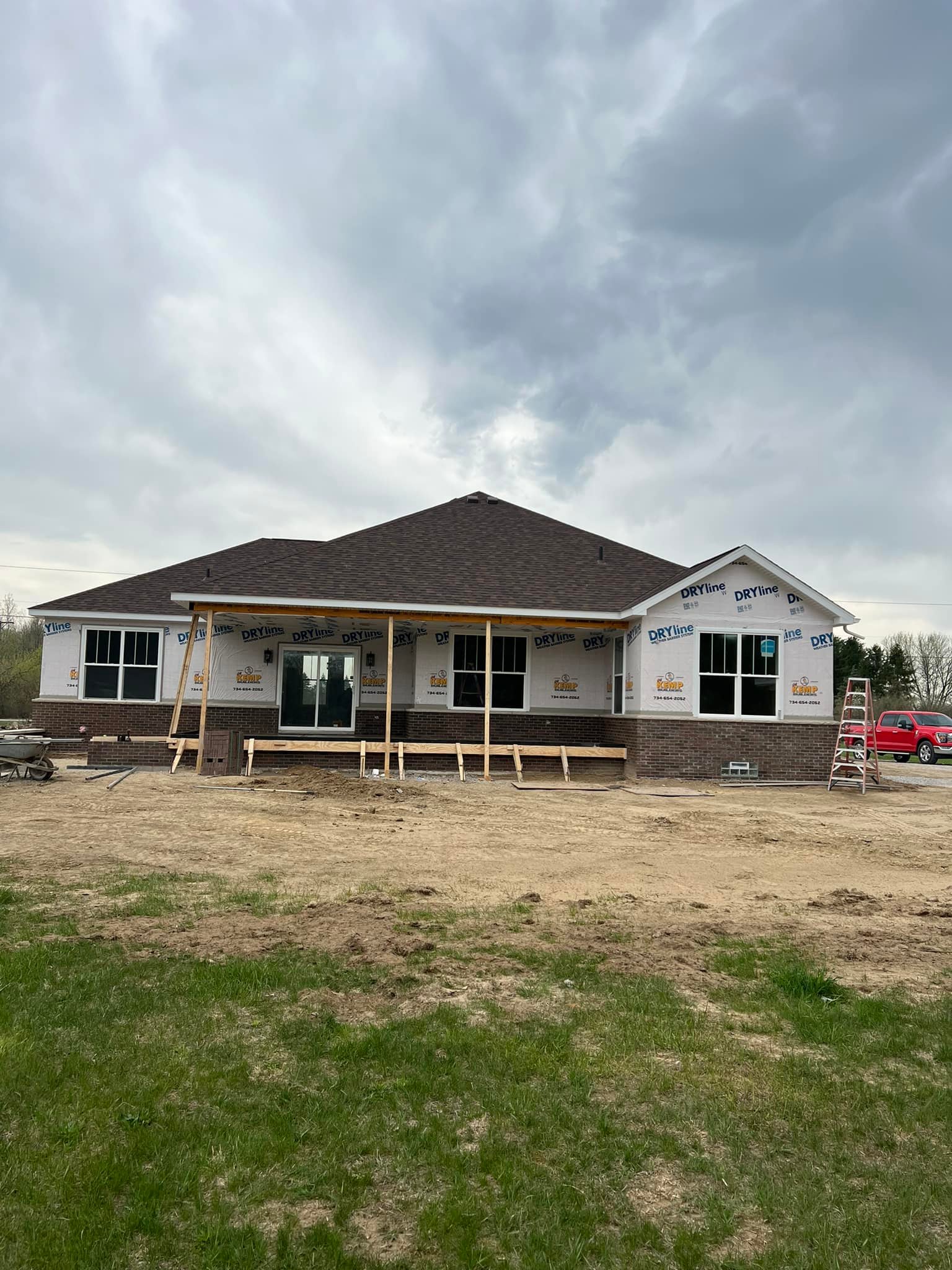 Partially constructed house with dark roof and brick base, wrapped in white building material. Overcast sky creates a calm, transitional atmosphere.