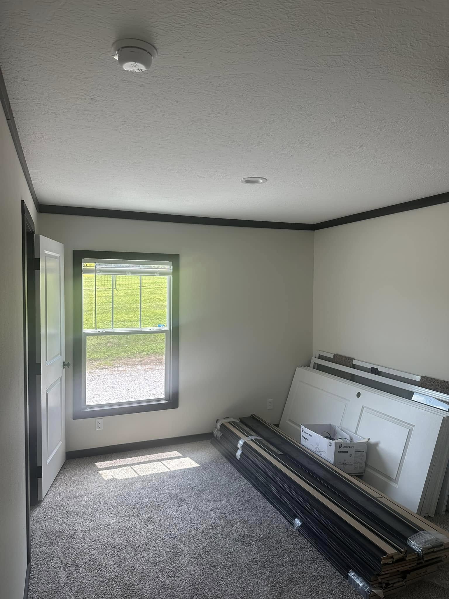 Small, unfurnished room with beige walls and gray carpeting. A window on the left lets in sunlight, illuminating stacked construction materials against the wall. Calm and minimal.
