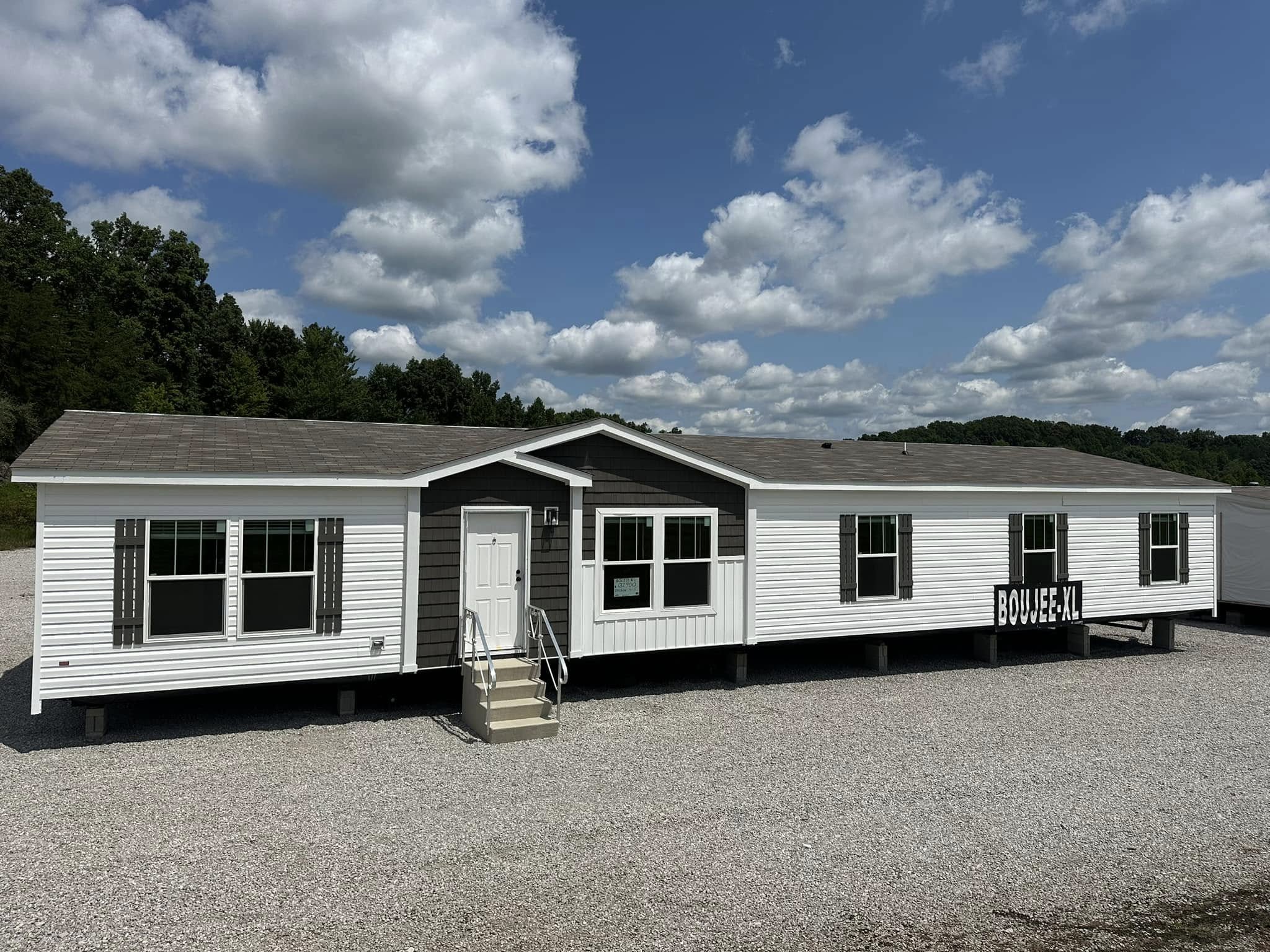 A white modular home labeled "Bouljee-XL" sits on a gravel lot under a partly cloudy blue sky. Trees are visible in the background, creating a serene atmosphere.