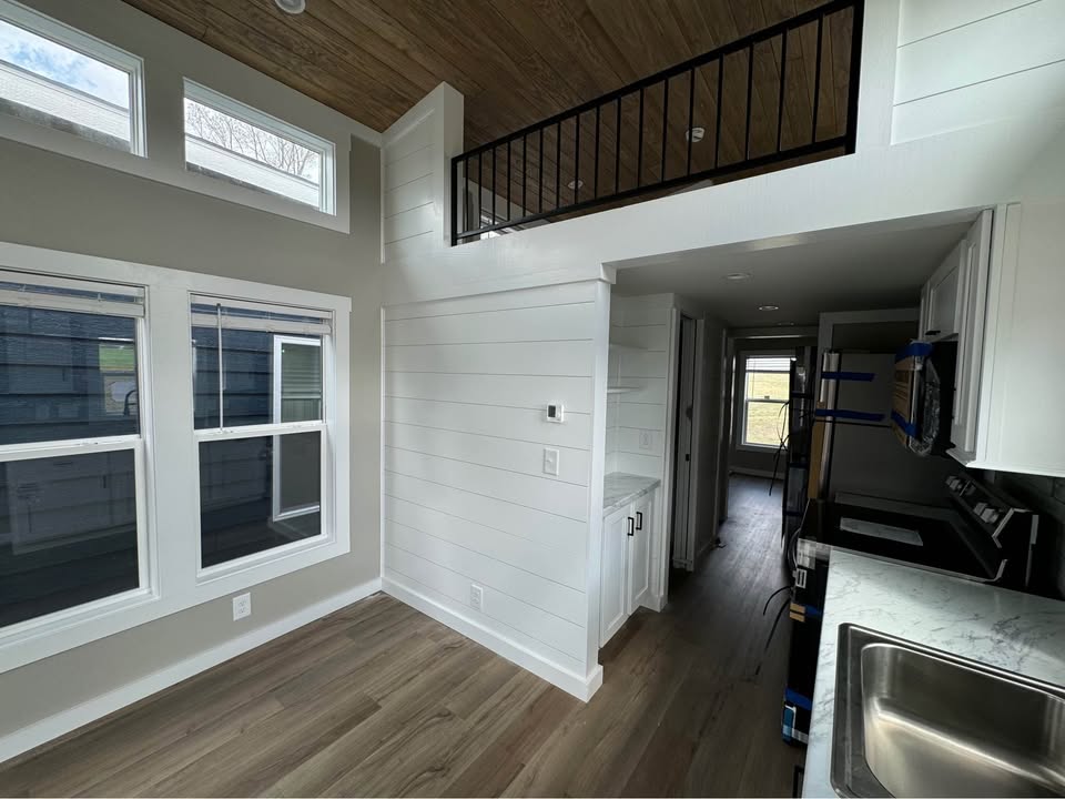 Modern tiny home interior with white walls, large windows, wood floors, and a small loft with a metal railing. Bright, open, and minimalist design.