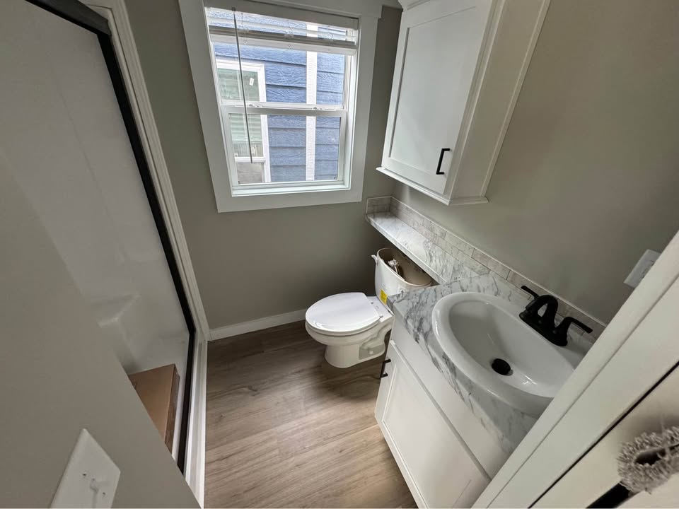 Compact bathroom with wood floor, gray walls, and white fixtures. Features a white toilet, marble countertop sink, and large window with natural light.