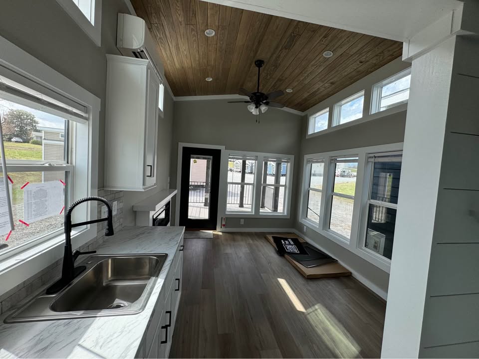 Compact modern kitchen and living area in a tiny home, featuring a wooden ceiling, sleek countertops, ample windows, and natural light. Cozy ambiance.