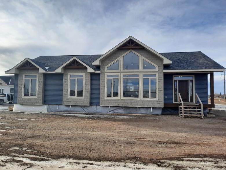 A blue single-story house with large geometric windows and a gabled roof sits on a dirt lot under a partly cloudy sky, creating a serene atmosphere.