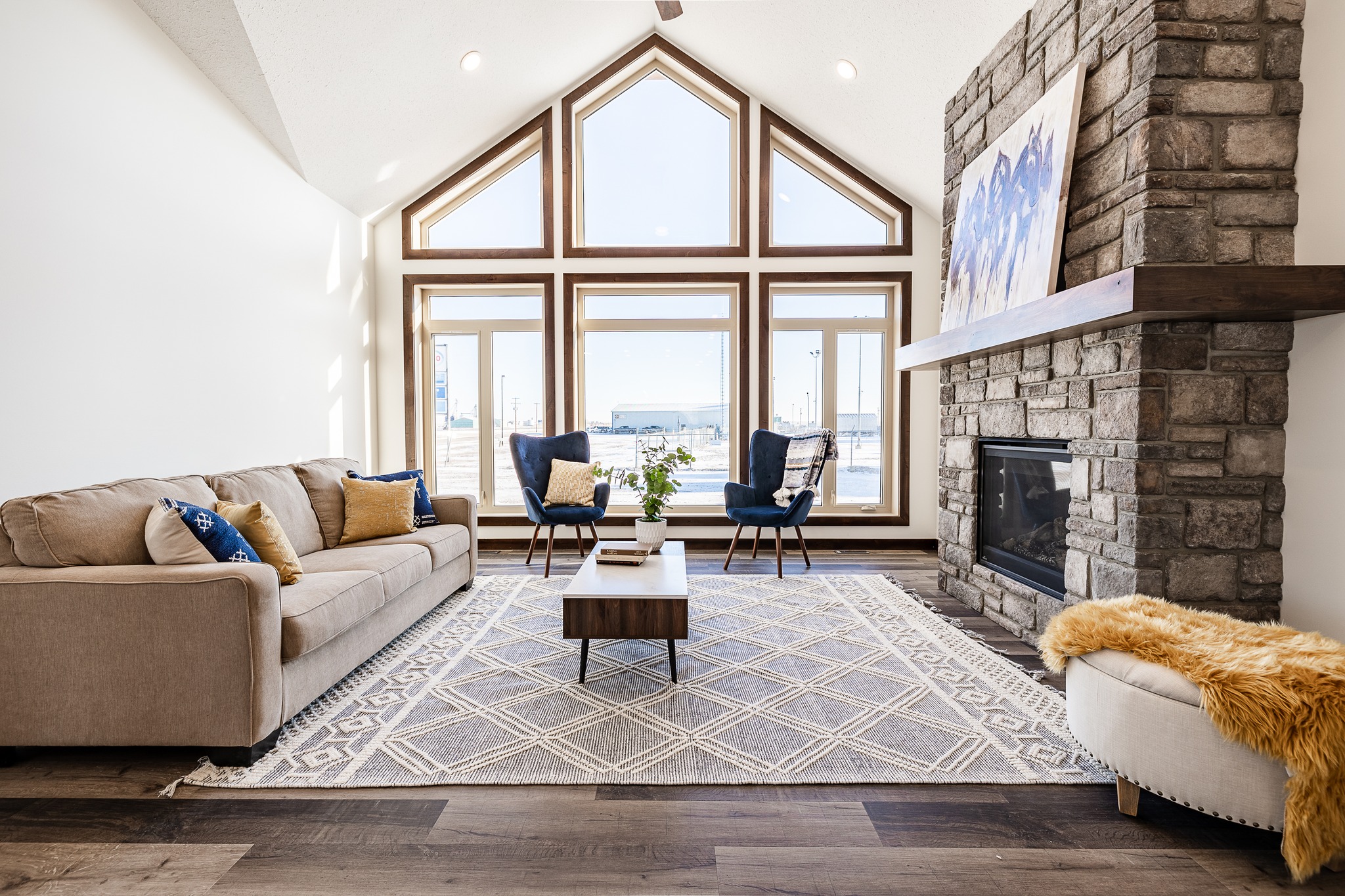 Spacious living room with vaulted ceiling, large windows, and stone fireplace. Cozy beige sofa, patterned rug, and modern decor create a tranquil atmosphere.