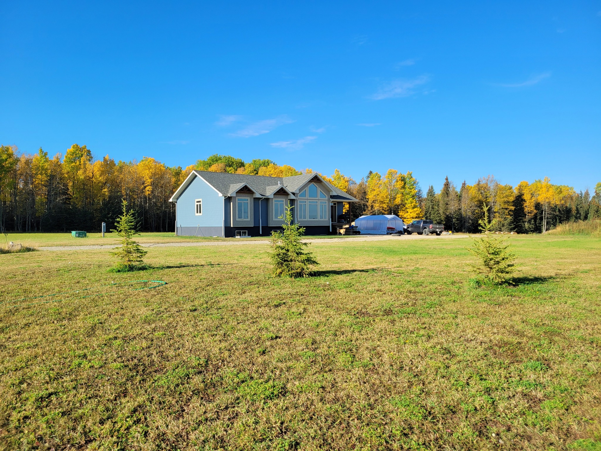 A blue house with large windows sits amid a vast green lawn. Small trees dot the front. In the background, a forest of autumnal trees under a clear blue sky.