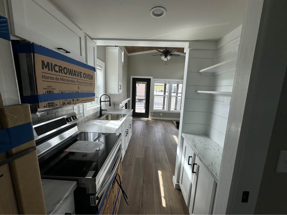 Modern, narrow kitchen with white cabinetry, marble countertops, and a boxed microwave. Wood floor leads to a bright living area with large windows.
