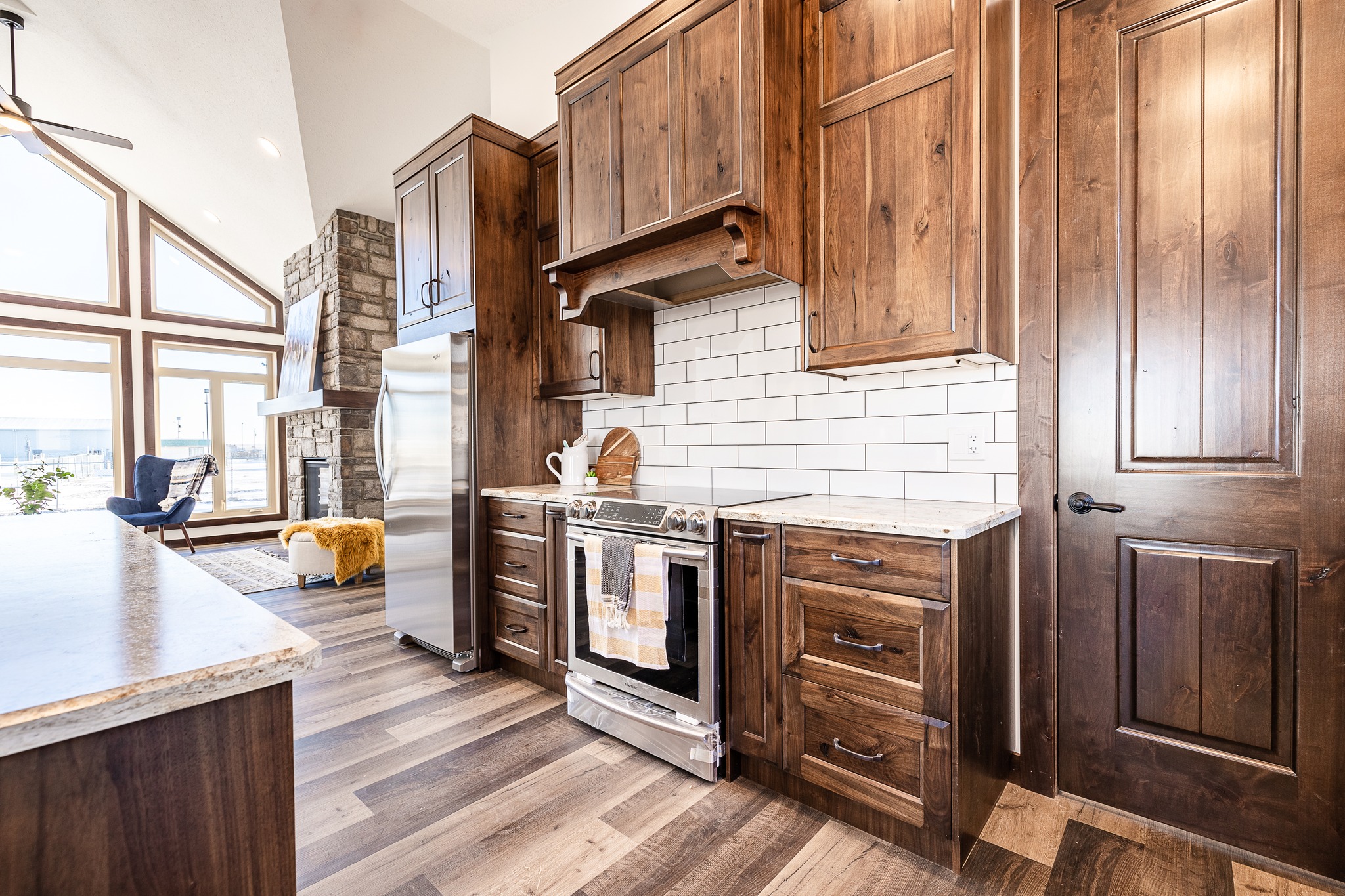 Spacious kitchen with rustic wooden cabinets, white subway tiles, and wood flooring. Bright, natural light streams through large windows, creating a warm, inviting atmosphere.