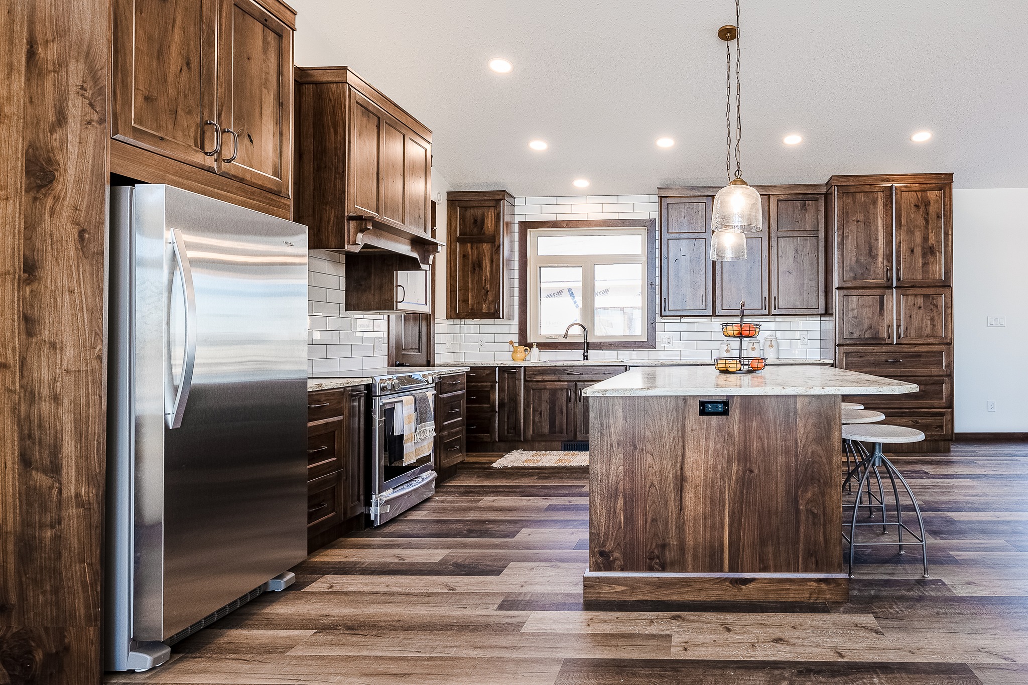 Spacious kitchen with rustic wooden cabinets, a large stainless steel refrigerator, and a central island. Bright pendant lights enhance the warm, inviting tone.