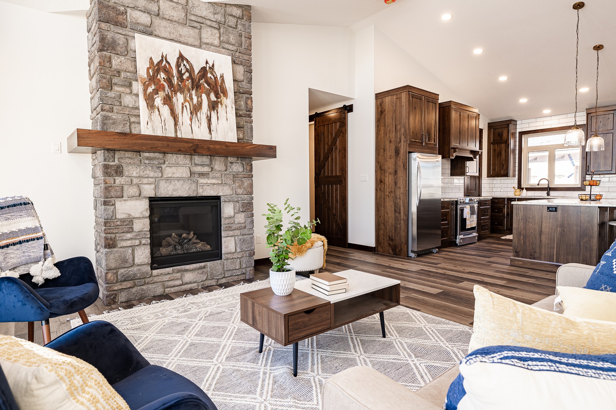 Cozy living room with stone fireplace and abstract art. Features navy chairs, wooden table with books and plant, open kitchen with pendant lights.