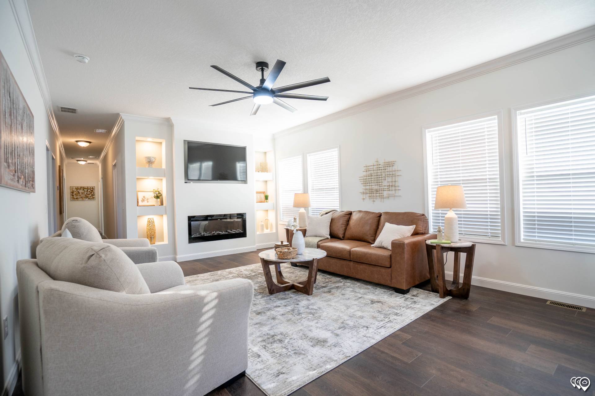 Modern living room with a brown leather sofa, two beige armchairs, and a patterned rug. A ceiling fan, wall-mounted TV, and fireplace add elegance. Elegant decor with bright ambiance.