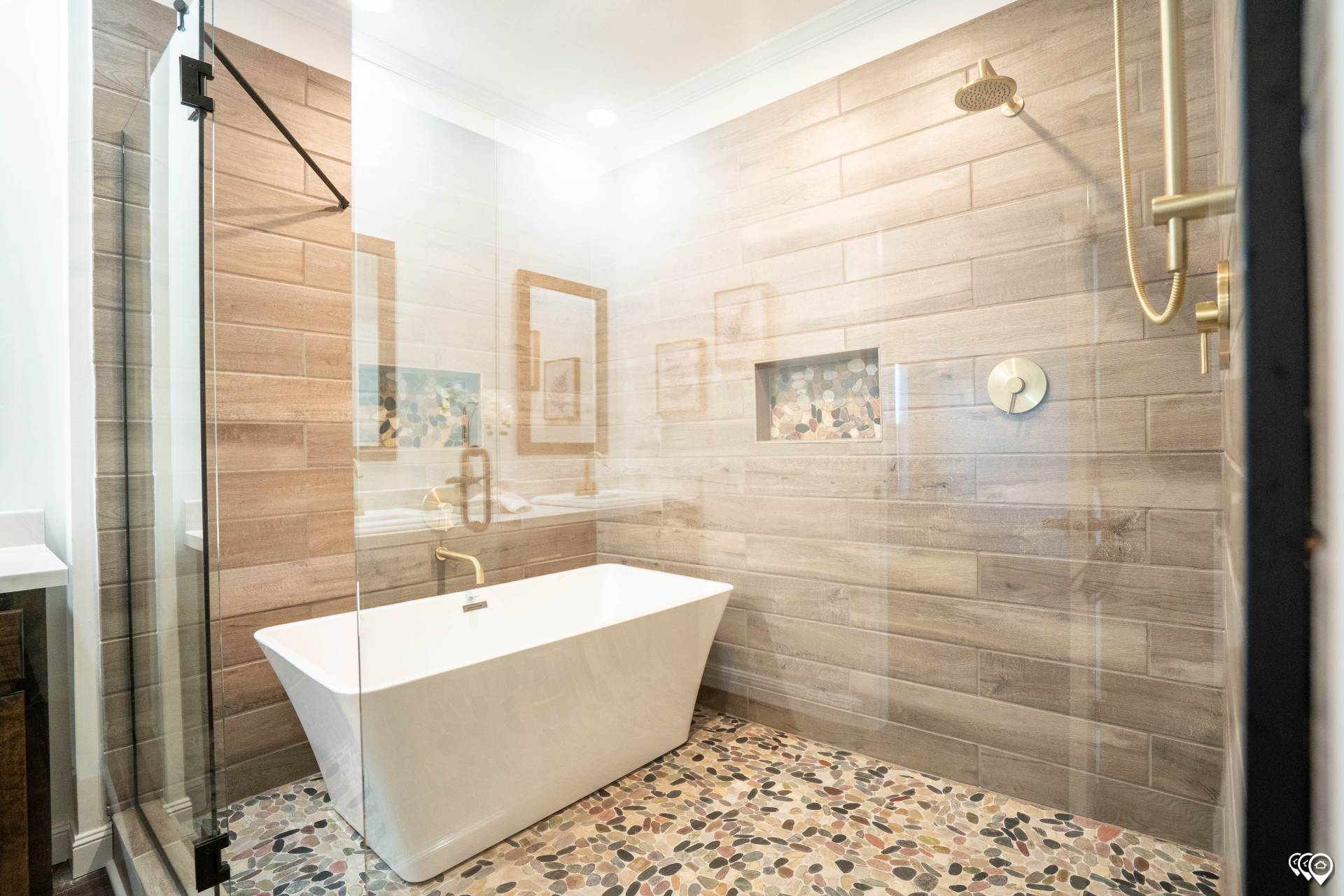 Modern bathroom with a freestanding white tub against a wood-paneled wall. Pebble-style floor, glass shower, brass fixtures, and warm ambient lighting.