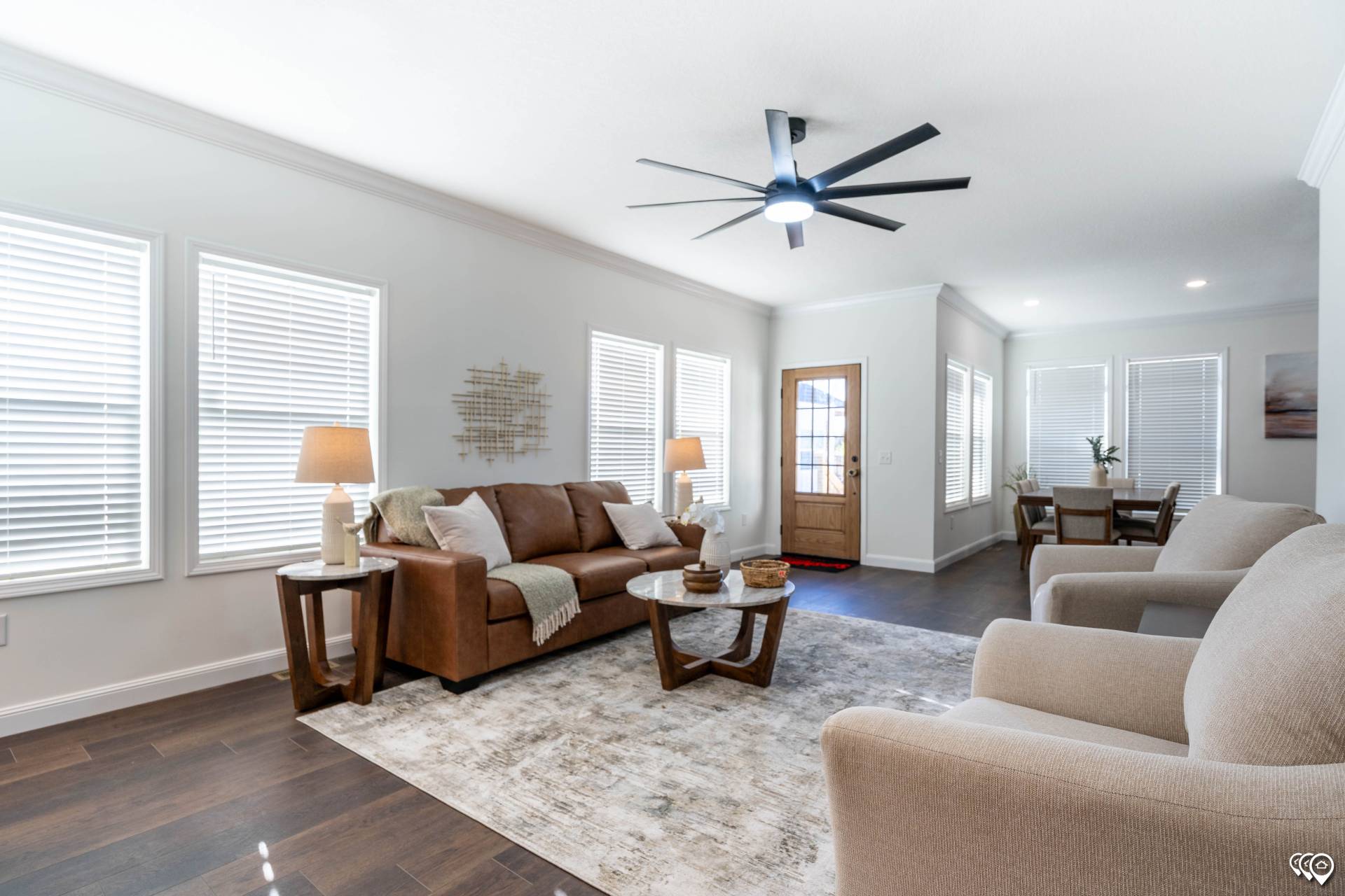Modern living room with a brown sofa, two beige armchairs, and wooden coffee table on a patterned rug. Bright natural light from large windows. Cozy and inviting.