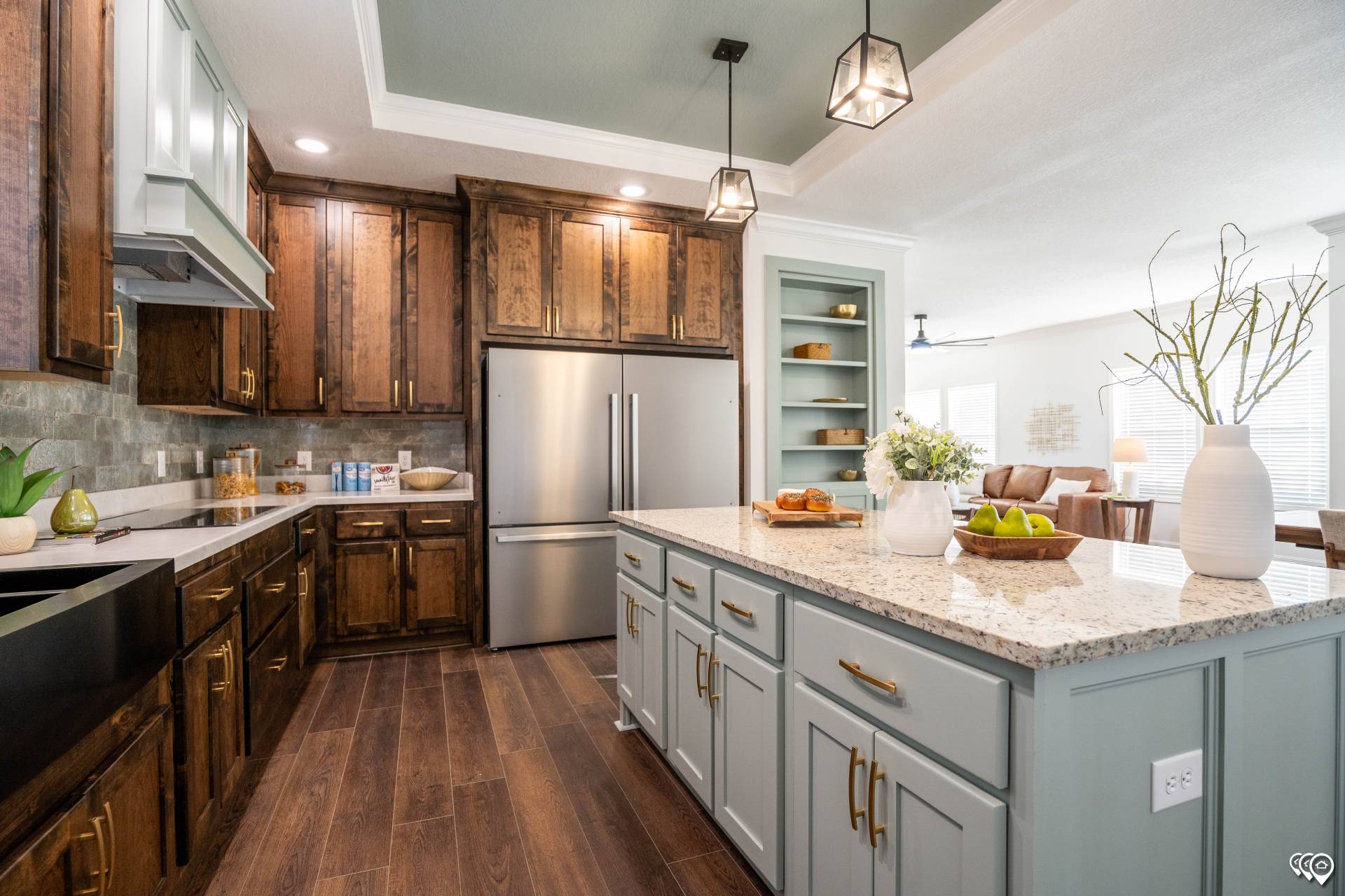 A modern kitchen with wood floors, dark wood cabinets, and a large island with a white granite countertop. Hanging pendant lights create a warm ambiance.
