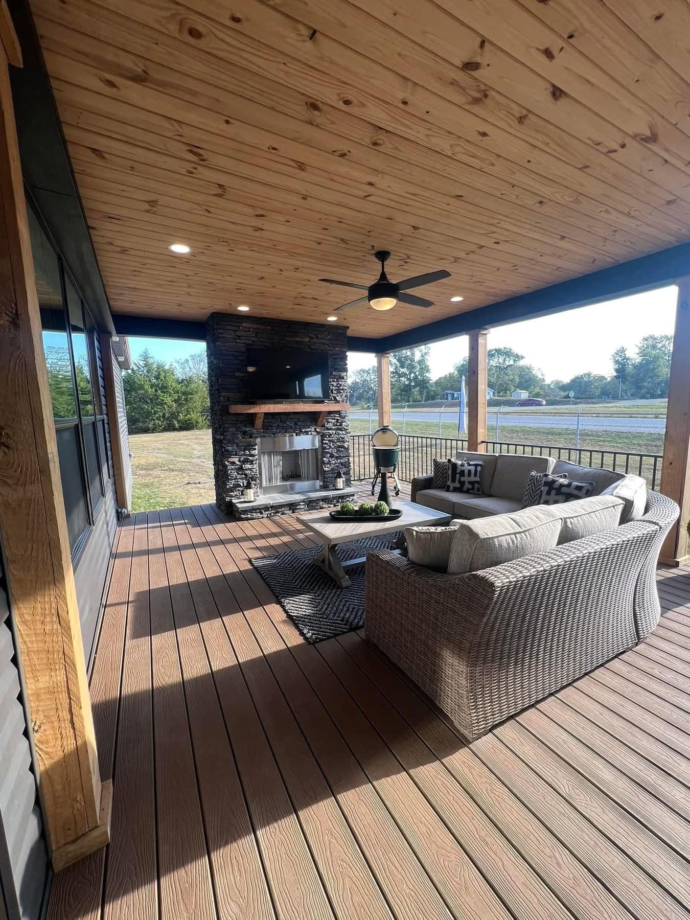 Covered patio with a wooden ceiling and deck, featuring a stone fireplace, outdoor sofa set, coffee table, and a ceiling fan. Peaceful and inviting. Image for illustrative purposes.