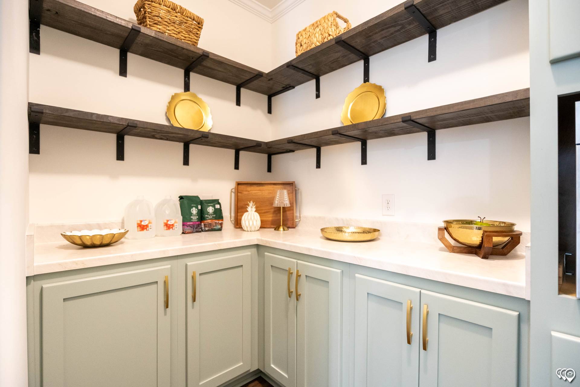 A cozy pantry with mint green cabinets and gold handles. Shelves hold wicker baskets and plates. Counters display a wooden cutting board and decorative jars.