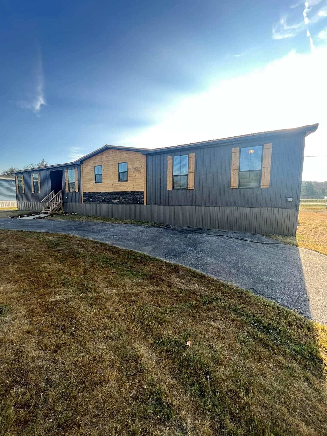 Single-story, modern mobile home with wooden accents and dark siding, set on a sunny day. Foreground features a grassy area and driveway. Image for illustrative purposes.