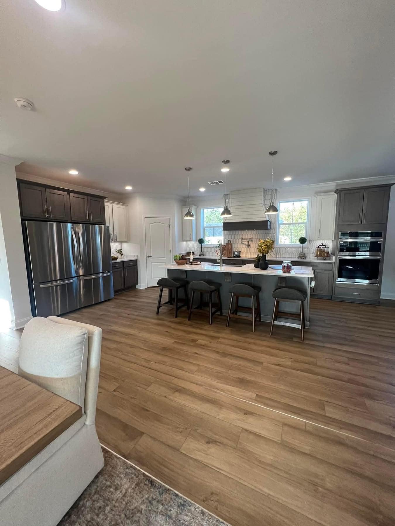 Spacious modern kitchen with wooden floors, a central island with four stools, stainless steel appliances, pendant lights, and large windows. Calm ambiance. Image for illustrative purposes.