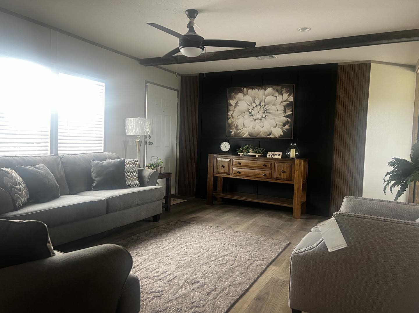 A cozy living room with a gray sofa and armchair, wooden console table, and large floral artwork. Soft lighting and a ceiling fan create a relaxing ambiance.