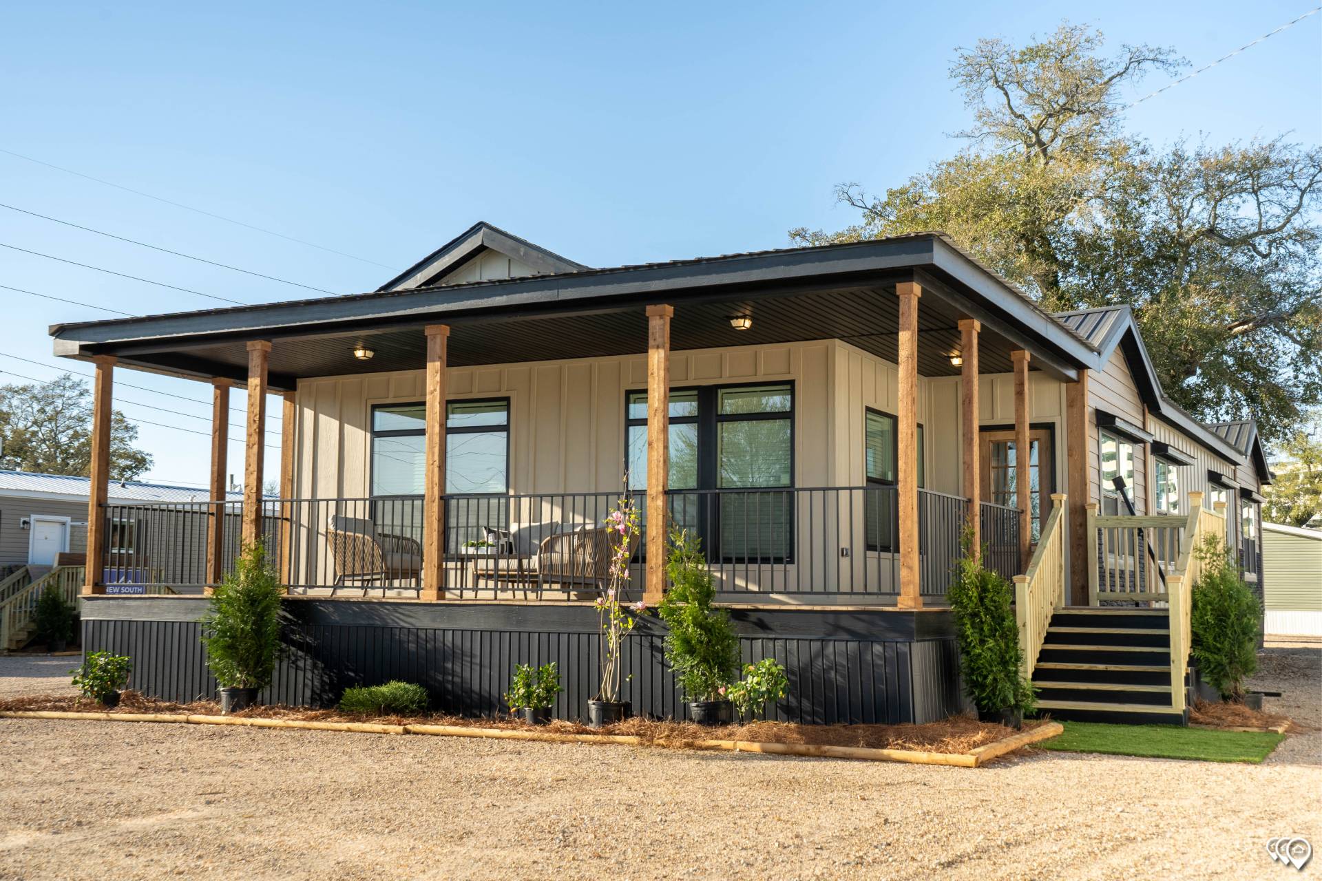 A modern tiny house with a wraparound porch, light beige siding, and dark metal roof. Wooden posts support the porch, surrounded by small green shrubs.