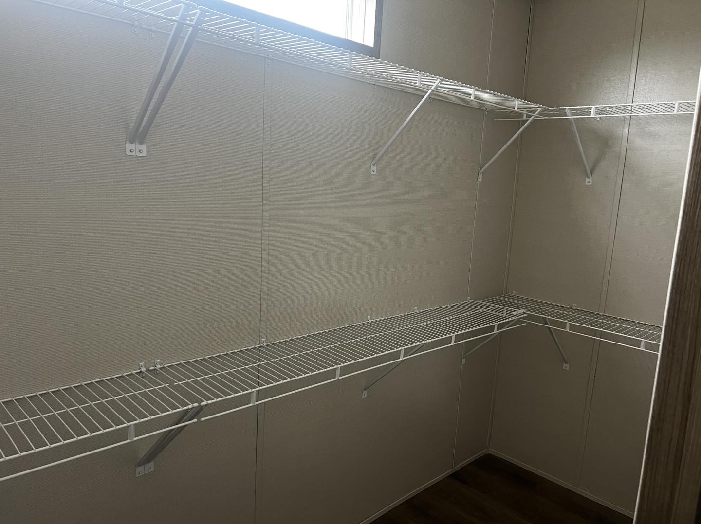 Empty closet with beige walls and wire shelving on two sides. Sunlight filters through a small window above, creating a clean and organized space.