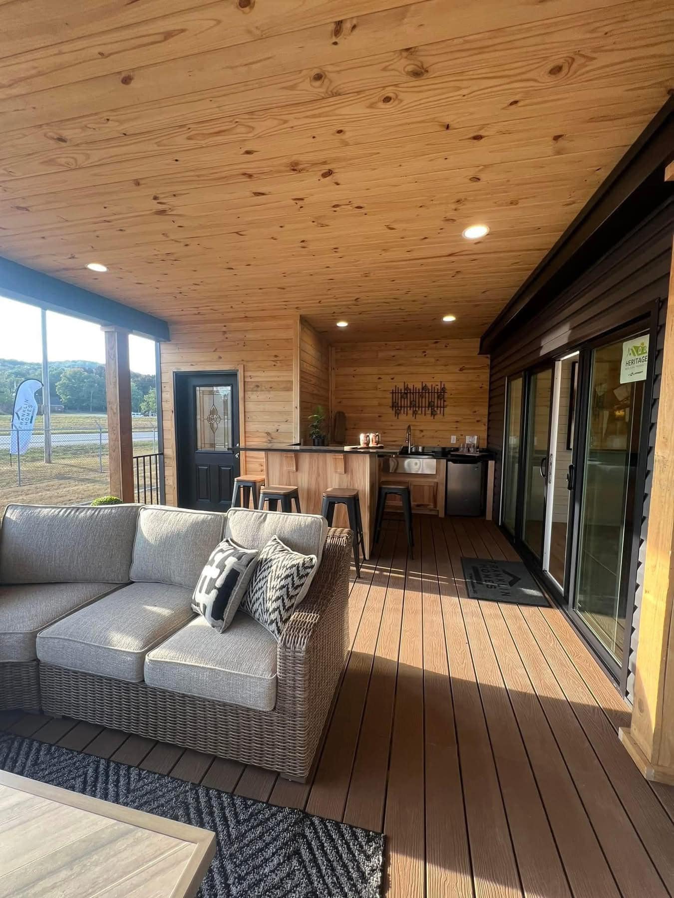 Stylish covered patio with wooden ceiling and deck, featuring a cozy wicker sofa with patterned cushions. Bar stools line a wooden counter next to glass doors. Image for illustrative purposes.