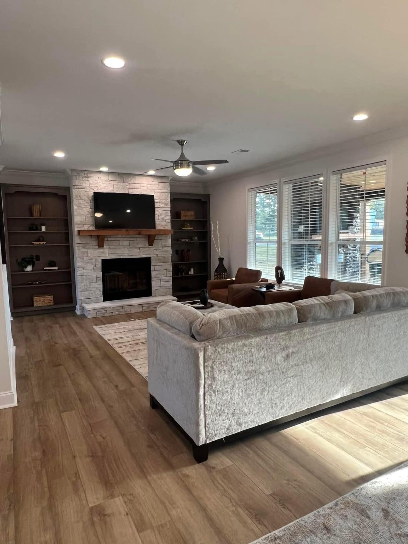 Spacious living room with cozy gray sofa, stone fireplace, wall-mounted TV, and wood shelves. Large windows brighten the warm, inviting space. Image for illustrative purposes.