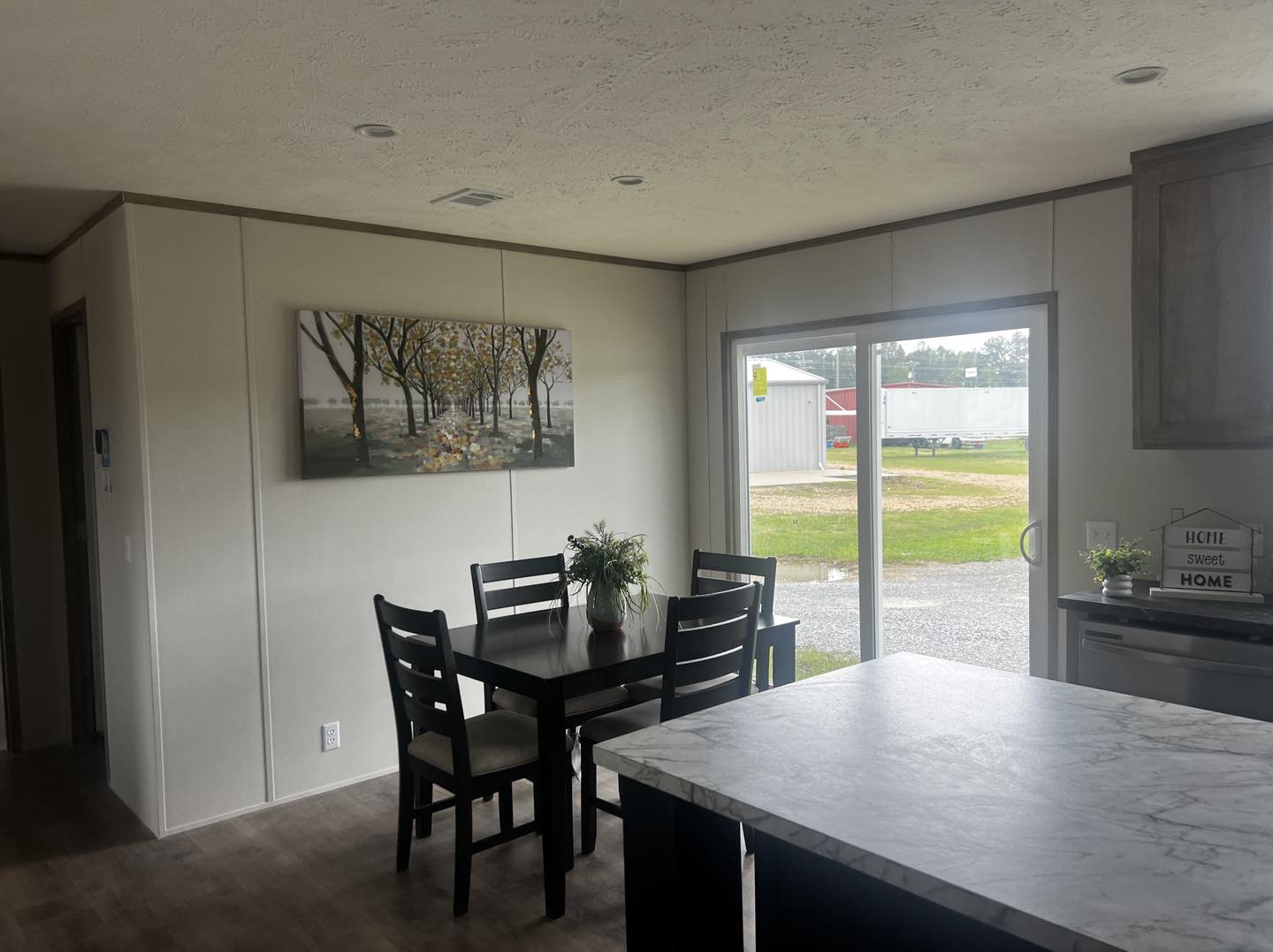 A cozy dining area with a dark wood table and chairs, a painting of trees on the wall, and a large window showing an outdoor view. A "Home Sweet Home" sign sits on the counter, adding warmth.