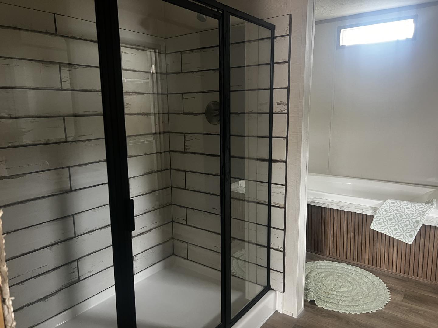 Modern bathroom with glass shower on the left, featuring white subway tiles and black frame. Bathtub on the right, white with wood paneling. Cozy, calm ambiance.