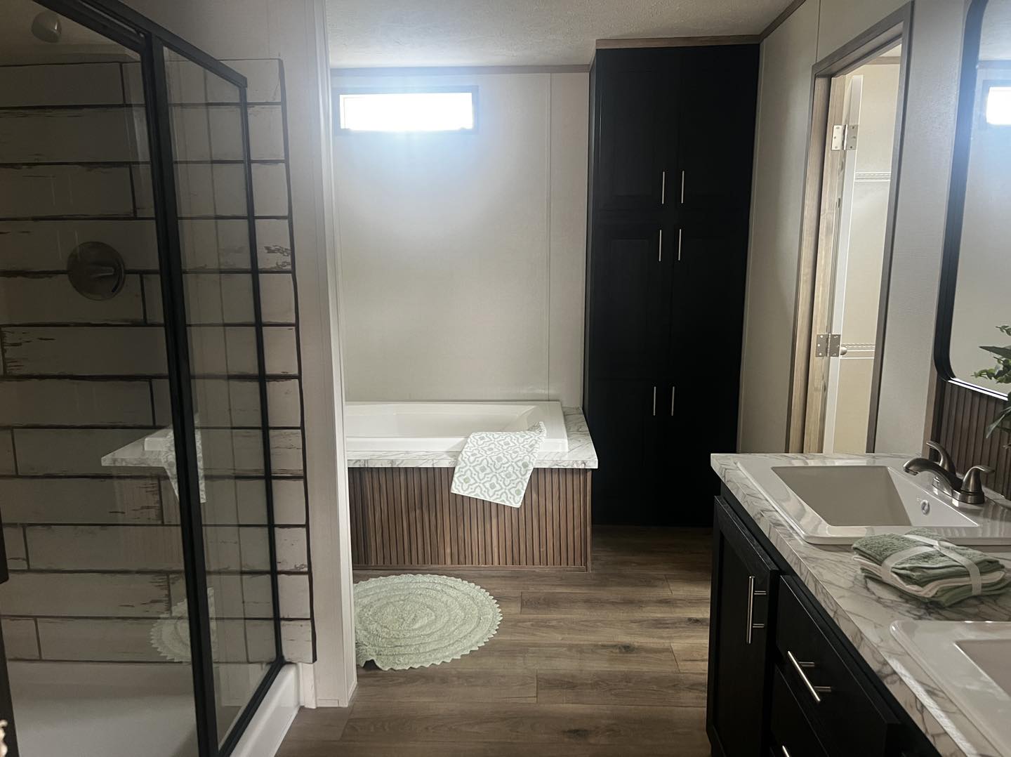 Modern bathroom featuring a glass-enclosed shower, a marble-top bathtub with a patterned towel, a dark wood double vanity, and a soft green rug.