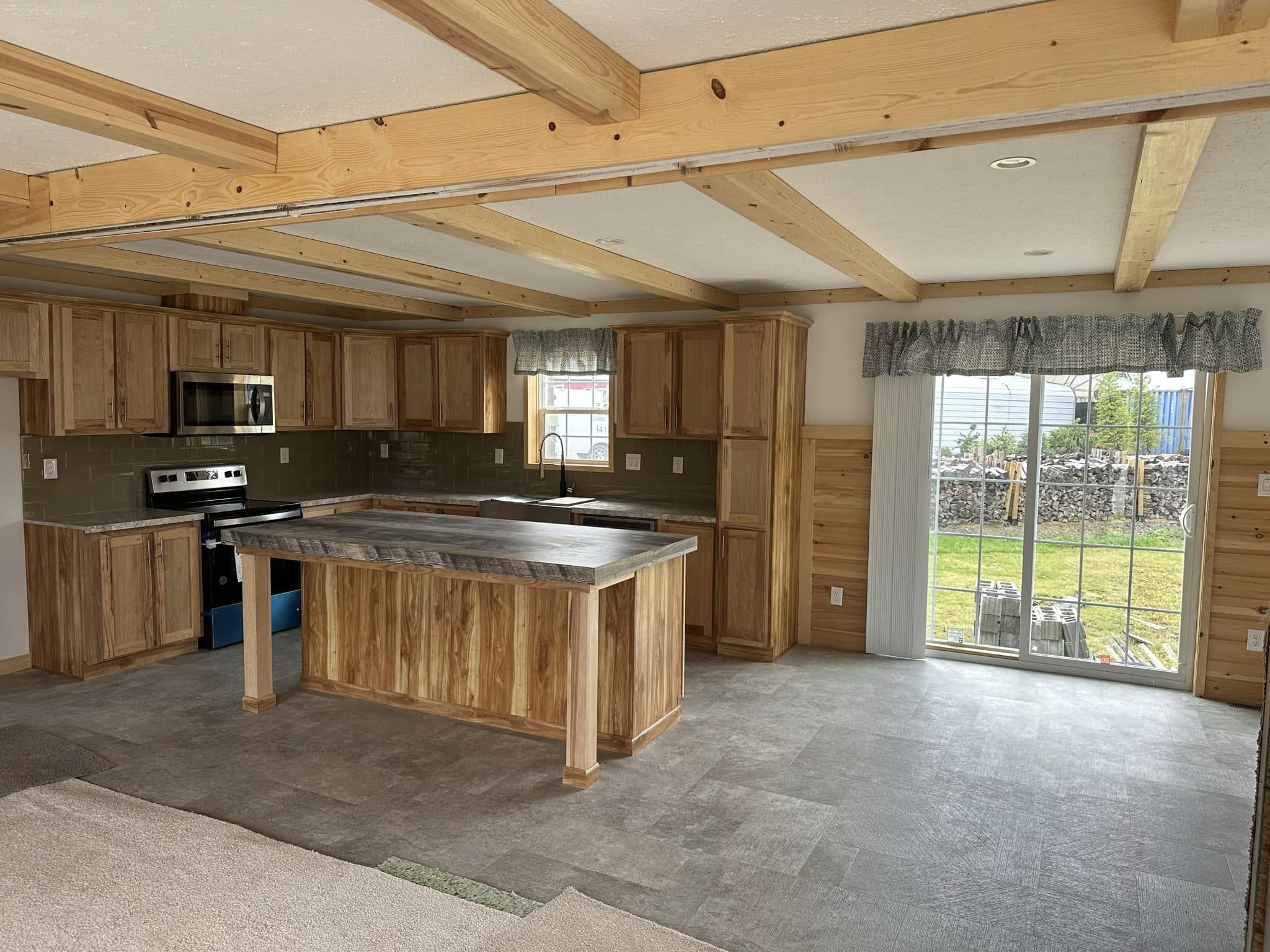 Spacious kitchen with wooden cabinetry, center island, and stainless steel appliances. Exposed beams on the ceiling. Sliding glass door leads to a grassy yard.