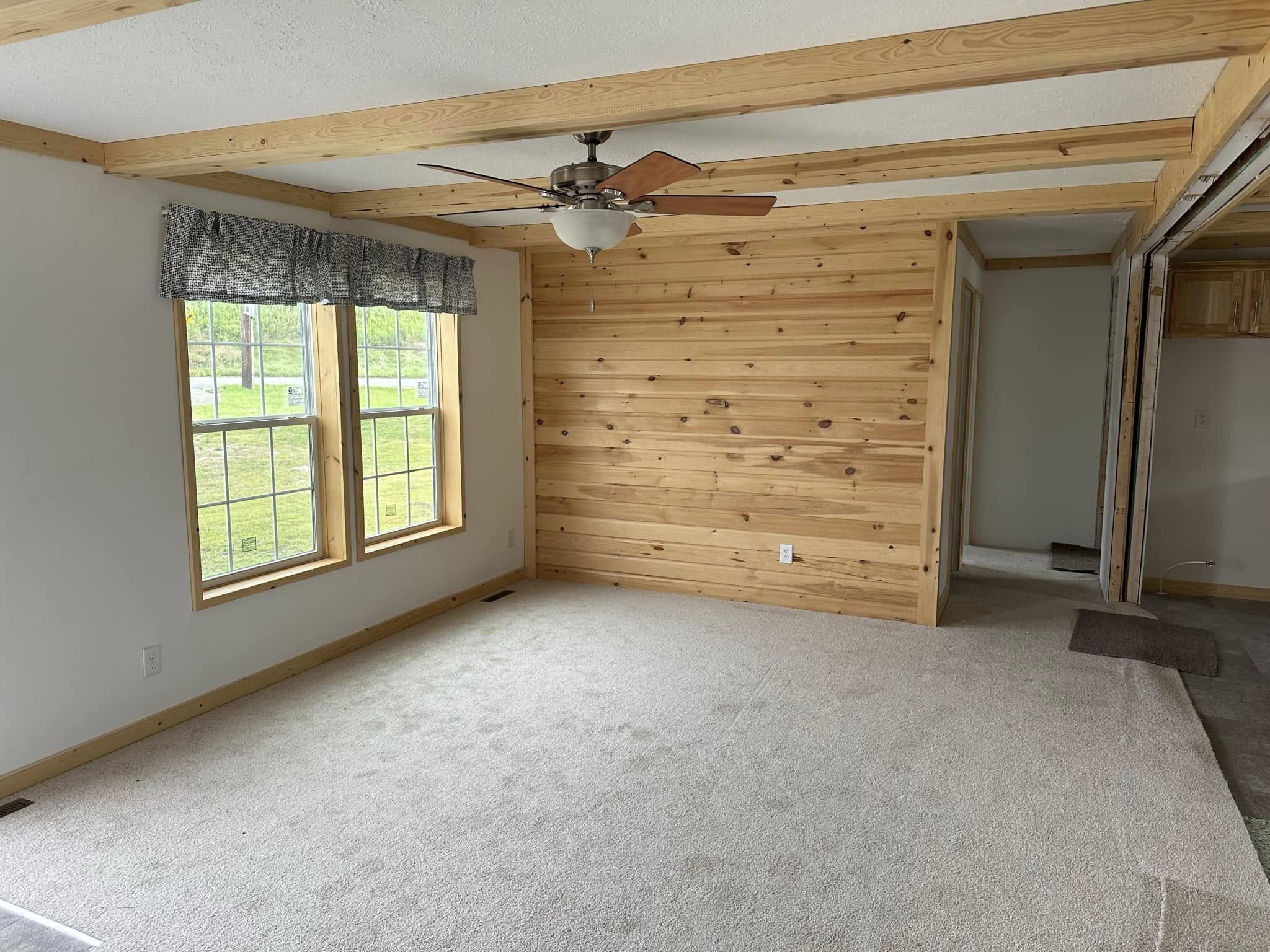 A cozy room with beige carpet, a wood-paneled wall, and exposed wooden beams. Two large windows with dark curtains let in natural light. A ceiling fan hangs centrally.