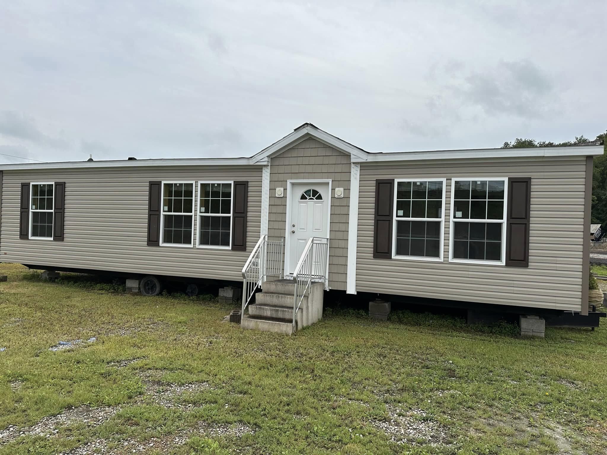 A beige manufactured home with a white front door, steps, and two large windows with shutters on each side, sits on a grassy area under a cloudy sky.