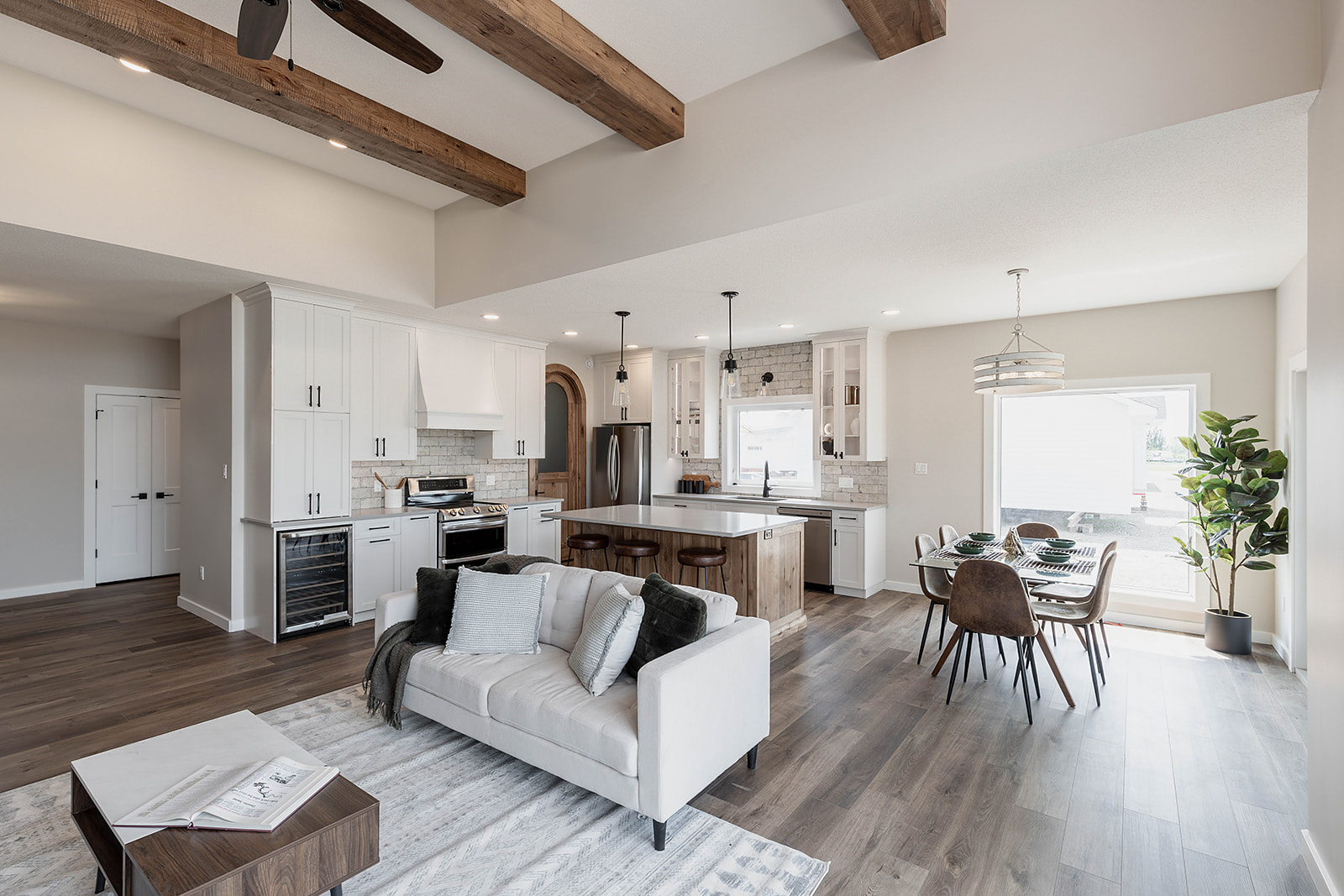 Spacious modern kitchen and living area with wooden beams, white cabinets, and a large island. Dining set near a bright window, plant decor adds warmth.