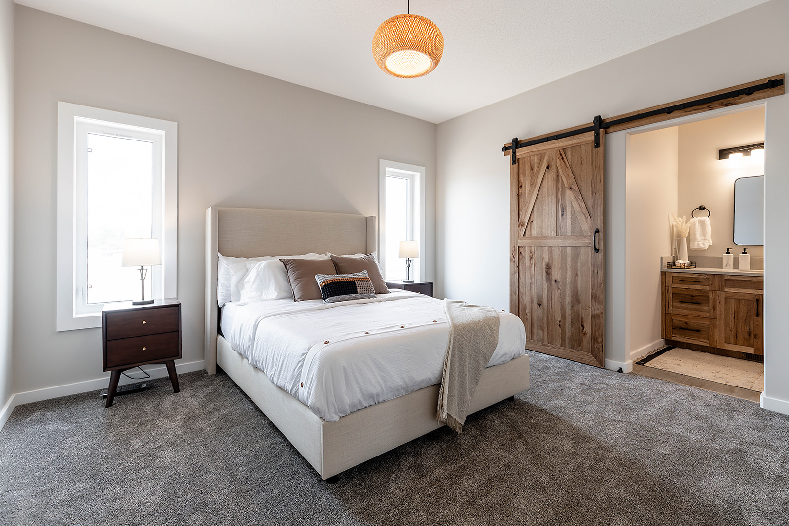 Cozy bedroom with a beige bed, soft covers, and pillows on a gray carpet. Warm lighting from a ceiling lamp and bedside lights. A wooden sliding barn door leads to a rustic bathroom, creating a serene, inviting ambiance.