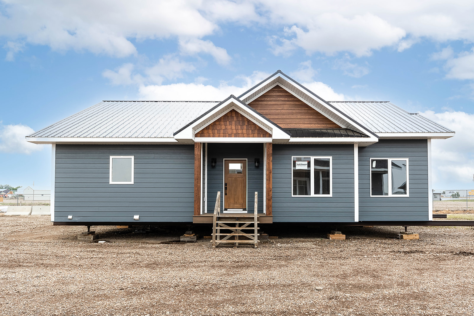 A modern modular house with blue siding, white trim, and a metal roof. It sits elevated on blocks with a wooden porch and steps, under a partly cloudy sky.