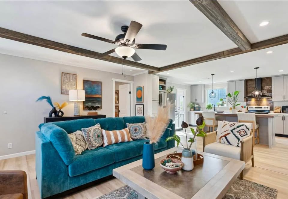 A cozy living room with a vibrant teal sofa adorned with patterned cushions, a modern coffee table, decorative plants, and a spacious, light-filled kitchen.