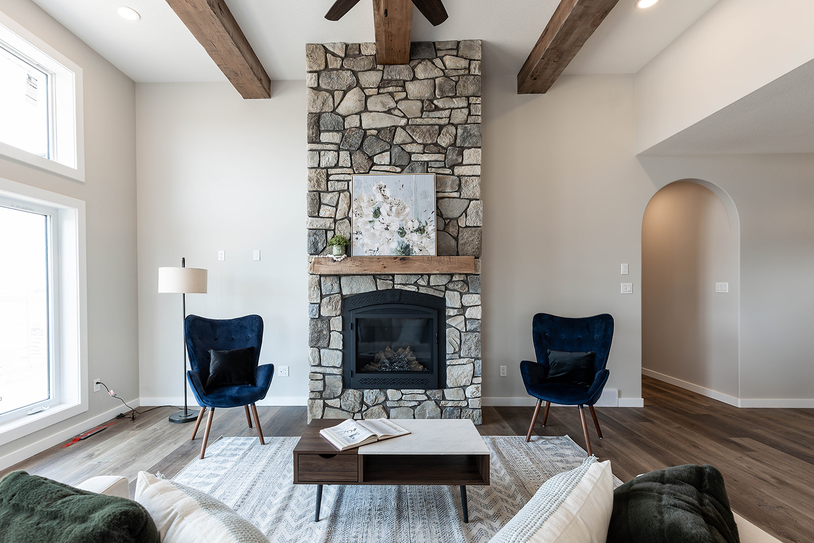 A cozy living room features a stone fireplace flanked by two blue chairs. Wood beams accent the ceiling, and large windows provide natural light.