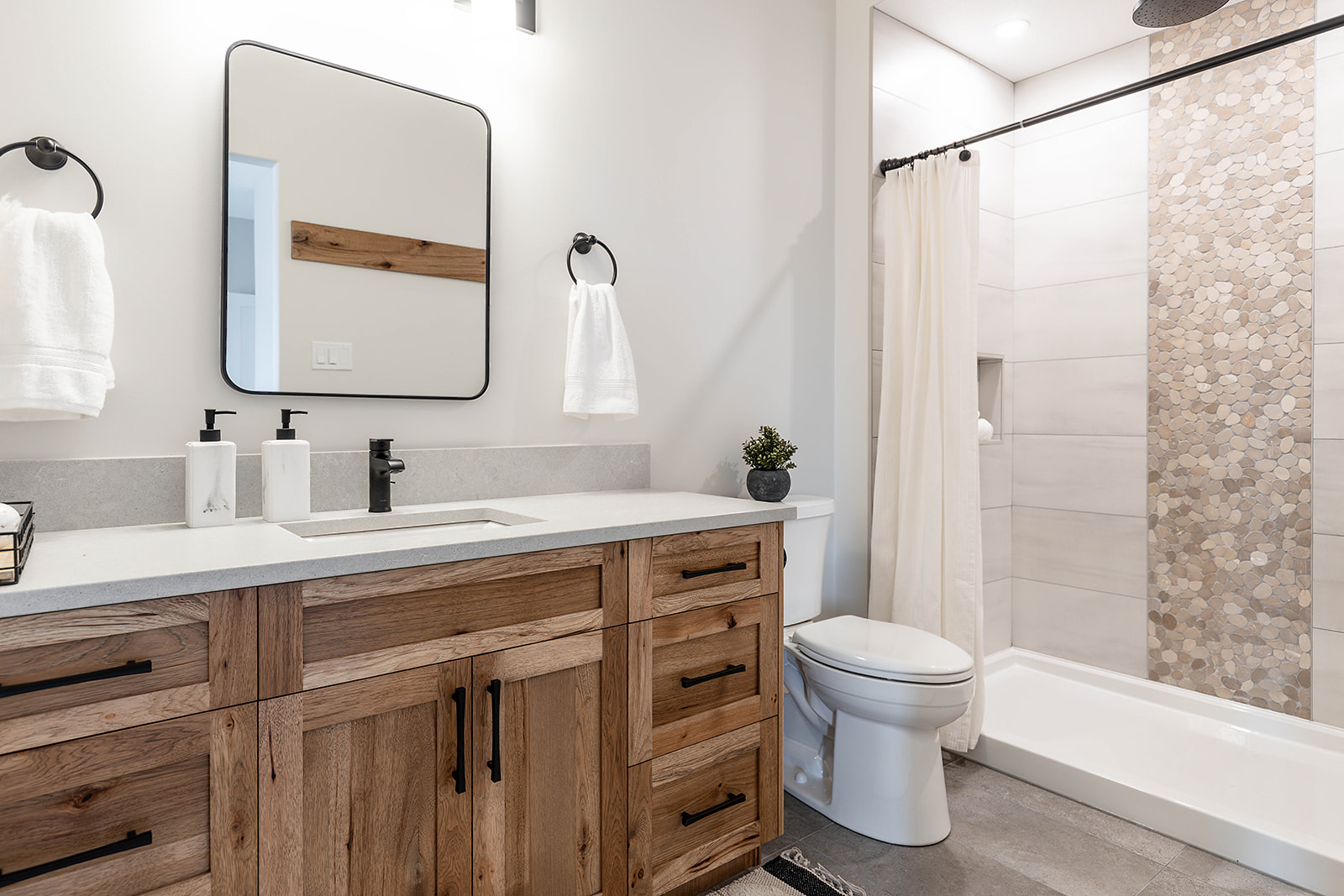 Modern bathroom with a wood vanity, black fixtures, and a rectangular mirror. Features a walk-in shower with mosaic tile accent and white curtain, creating a warm, minimalist feel.