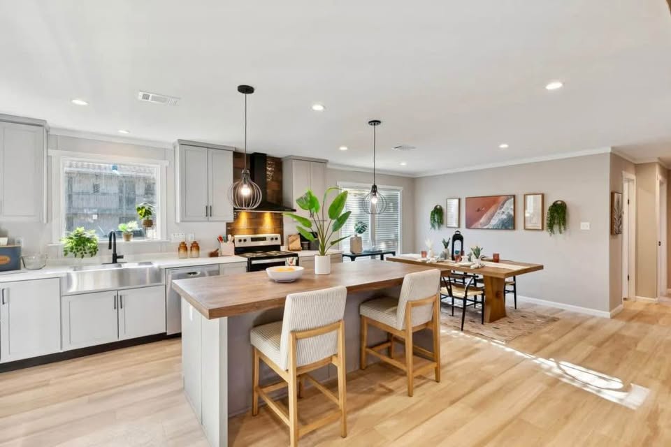 Bright, modern kitchen and dining area with light wood flooring. Features a wooden island with stools, gray cabinets, plants, and stylish pendant lights.