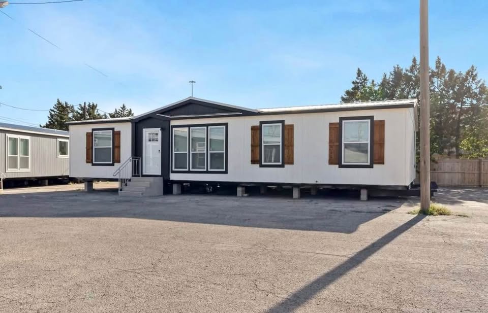 A white manufactured home with brown shutters is set on a paved lot. It has a small staircase leading to the door. Trees and a fence are in the background.