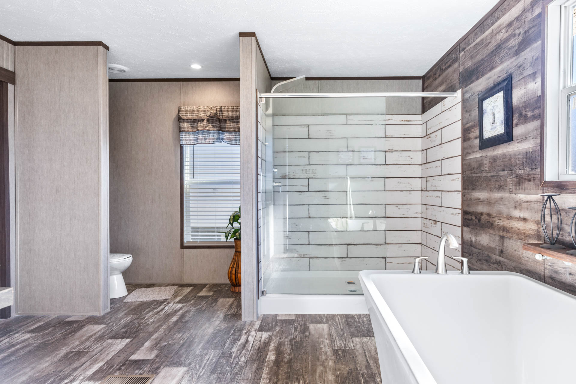 Modern bathroom with wood-patterned tile floor, walk-in shower, and freestanding tub. Large window and rustic decor create a cozy, airy feel.