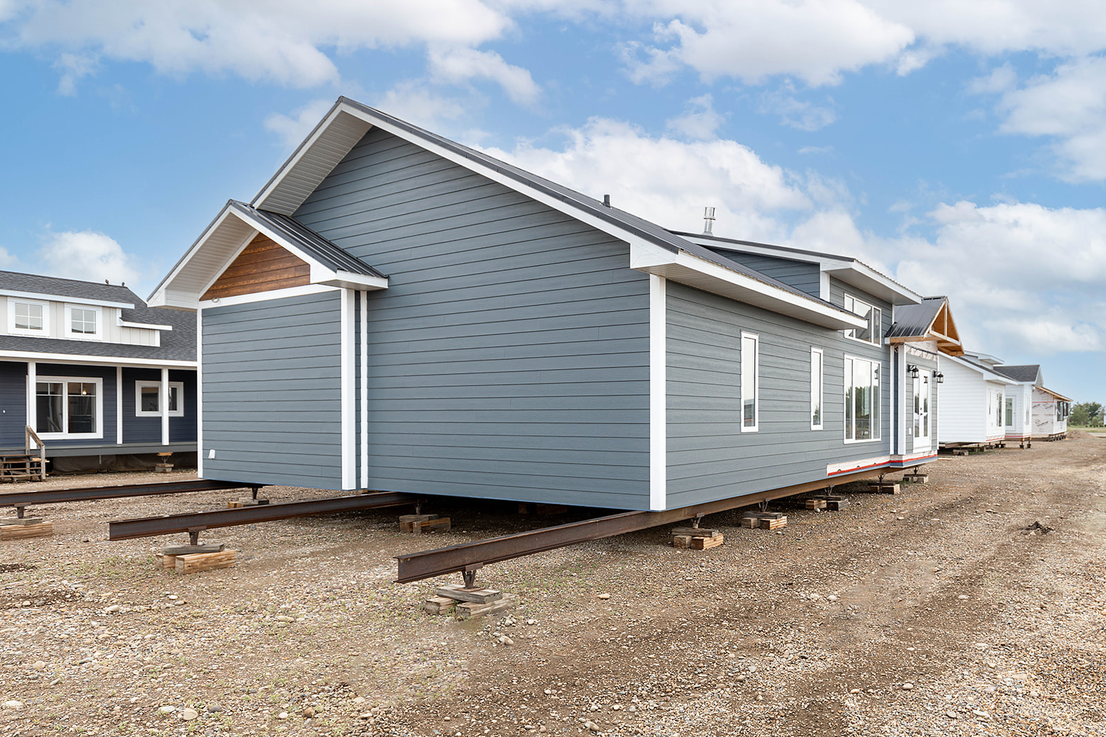 A blue modular home with white trim sits elevated on beams against a clear sky. Several similar homes are aligned behind it on a dirt lot.