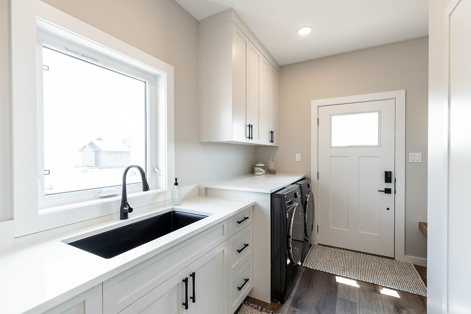 Bright laundry room with beige walls, white cabinetry, and laminate flooring. Features a black washer and dryer, a black sink, and a window by the door.