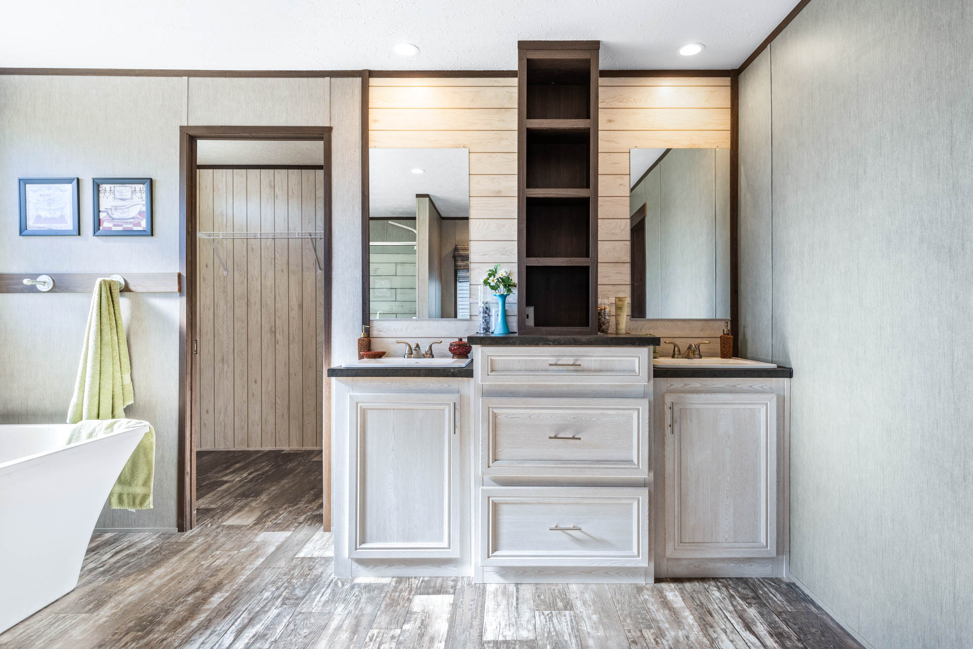 Modern bathroom with dual sinks and wooden cabinets, framed by two mirrors. A green towel hangs nearby. The floor has a wood-like finish.