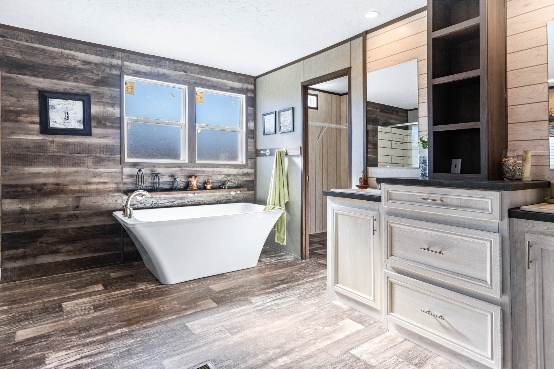 Spacious bathroom with a freestanding white bathtub against a rustic wooden wall, two windows, green towel, and light wood cabinets. Calm, inviting ambiance.